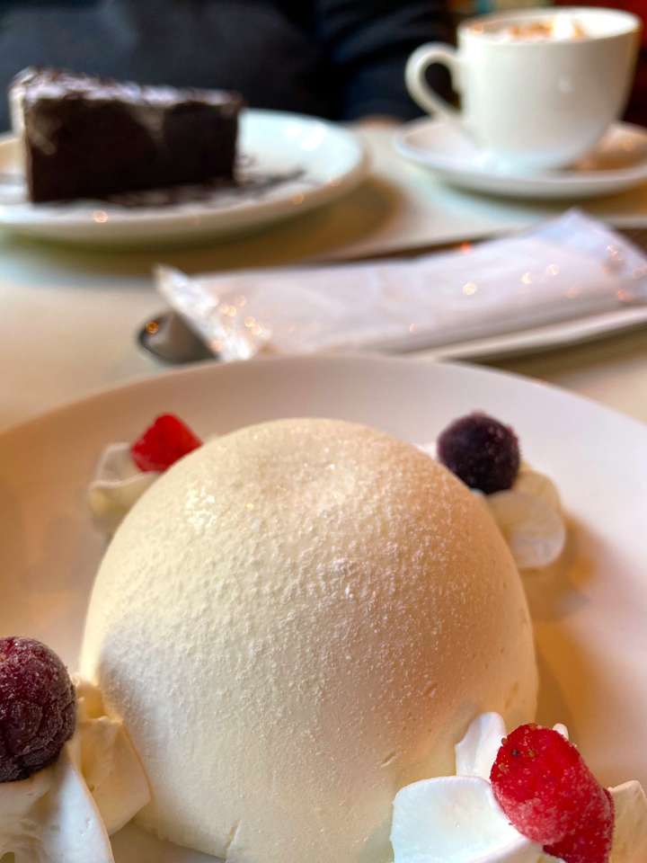 A close-up of a dome shaped unbaked cheesecake garnished with whipped cream and berries on a plate. In the background, there is a slice of chocolate cake and a cup of coffee on the table.