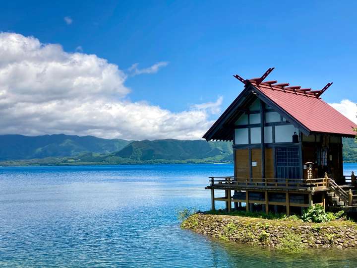 A small wooden structure with a red roof stands on a stone foundation over clear blue water, against a backdrop of green hills and a partly cloudy sky. The scene is serene, with vibrant colours of nature under bright daylight.